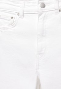 Gros plan sur un jean en denim blanc montrant la poche avant, le bouton en métal, les passants de ceinture et les détails des coutures.