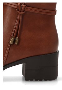 Botín de cuero marrón hasta el tobillo con punta redonda, tacón negro plano, cordones decorativos y adornos metálicos. Textura suave con un acabado mate.