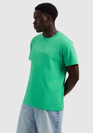 Man in een groen T-shirt met korte mouwen met de tekst "PurePath", lichtblauwe spijkerbroek, kijkt naar links, handen achter de rug, neutrale grijze achtergrond.