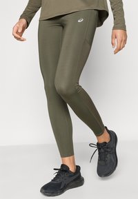 Κολάν σε στενή εφαρμογή, ελιάς χρώματος, με λεία υφή και αντίθετες ραφές, συνδυασμένα με μαύρα αθλητικά παπούτσια.