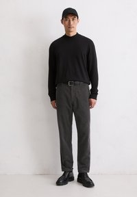 Schwarzes Langarmshirt kombiniert mit gestreifter grauer Cordhose. Ein schwarzer Gürtel und eine Kappe vervollständigen das Outfit. Schwarze Schnürschuhe.