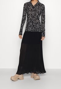Blouse noire à manches longues avec des pois blancs, associée à une jupe maxi plissée noire et des chaussures à plateforme beiges. Les textures des tissus sont lisses.