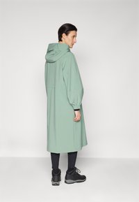 Manteau vert, à capuche, de longueur mi-longue avec une coupe décontractée, des poignets élastiques et sans fermetures visibles, associé à un pantalon noir et des bottes noires robustes.