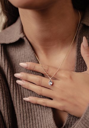 Gold necklace featuring a teardrop-shaped pendant with a blue gemstone, worn on a person’s neck. Knit sweater texture visible in background.
