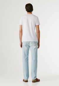 Mann trägt ein schlichtes weißes T-Shirt mit kurzen Ärmeln, hellblaue Jeans und braune Schuhe, steht mit dem Rücken zum Betrachter auf weißem Hintergrund.