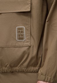 Gros plan sur une poche de veste marron avec un patch rectangulaire cousu présentant un logo de crocodile et un ourlet et poignet élastiqués.