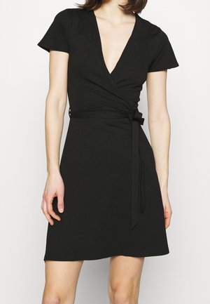 Frau trägt ein kurzes schwarzes Wickelkleid mit V-Ausschnitt und gebundenem Taillengürtel, steht vor einem schlichten hellen Hintergrund.
