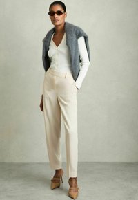Cardigan bianco a coste con bottoni, scialle grigio lavorato a maglia, pantaloni beige sartoriali e scarpe con tacco color nocciola con dettagli a cinturino. Inclusi occhiali da sole.