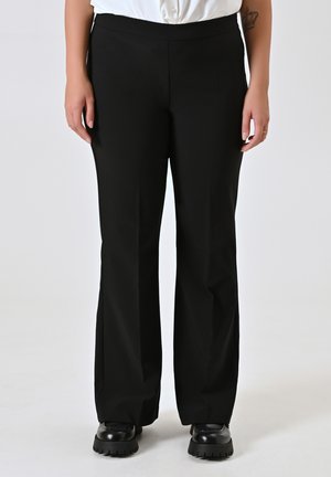 Pantalons noirs ajustés avec une texture lisse, une légère évasement à l'ourlet et une taille large. Portés avec des chaussures noires.