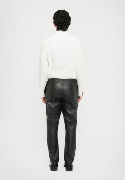 Man standing facing away, wearing a white long-sleeve shirt, black textured leather pants, and black shoes against a plain background.