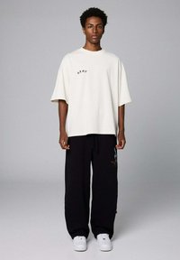 Cremfarbens Oversized-T-Shirt mit besticktem Text, kombiniert mit schwarzen weit geschnittenen Hosen mit grafischem Design und weißen Sneakern.