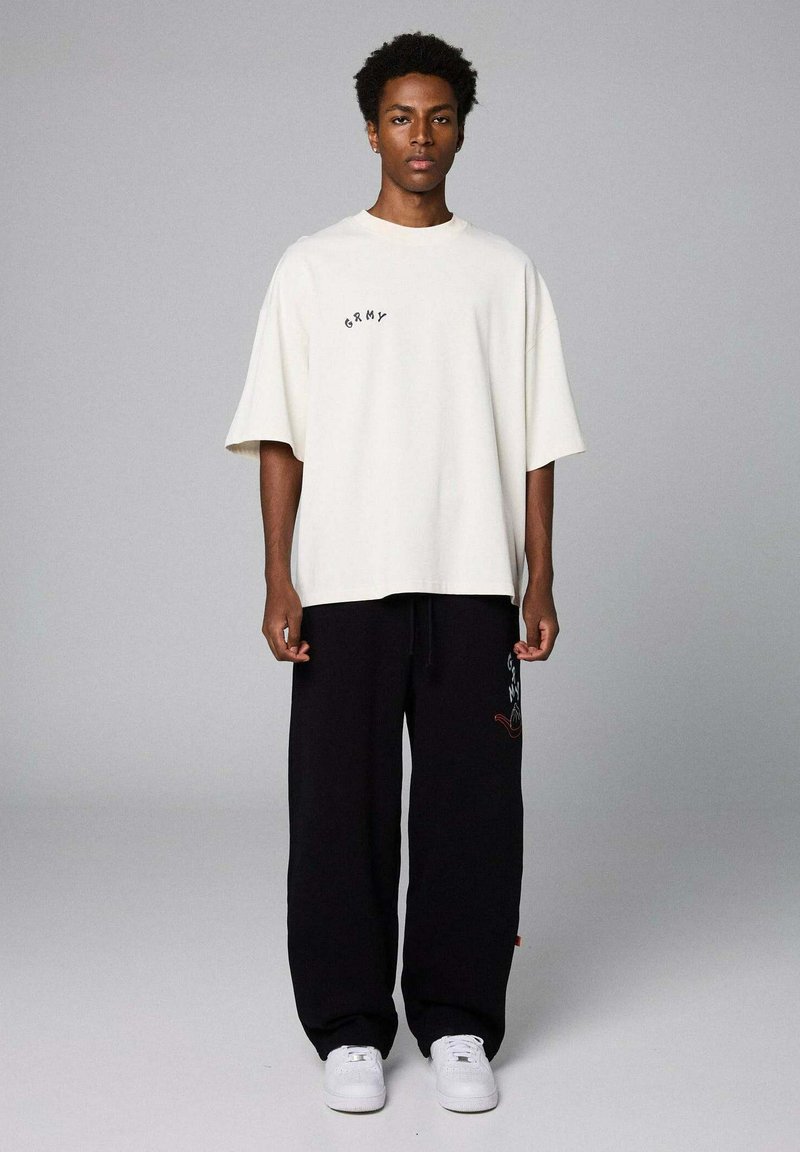 Cremfarbens Oversized-T-Shirt mit besticktem Text, kombiniert mit schwarzen weit geschnittenen Hosen mit grafischem Design und weißen Sneakern.