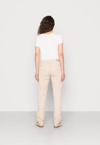 Calças beiges com um design reto, apresentando bolsos traseiros e um rótulo de couro castanho na cintura. Combinadas com uma t-shirt branca simples.