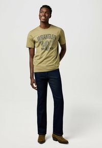 T-shirt vert olive avec un graphisme "WRANGLER 1947", associé à un jean bleu foncé et des bottes en suede marron. Le modèle se tient dans une pose décontractée.