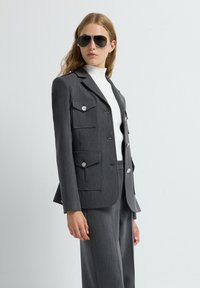 Blazer gris entallado con dos bolsillos delanteros y detalles en botones plateados, combinado con pantalones grises y un suéter de cuello alto blanco.