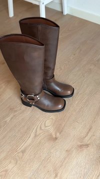 Bottes en cuir marron jusqu'aux genoux avec une finition lisse, dotées d'une boucle et d'un détail de sangle en ton argent. Bout carré et talon bas.