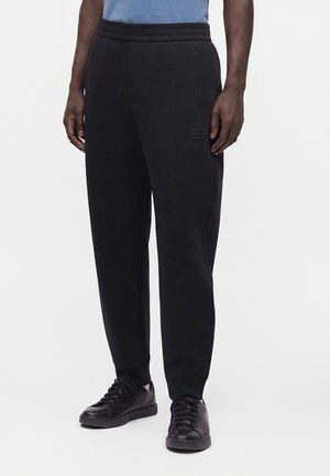 Homme portant un pantalon de jogging noir et des baskets noires, debout les mains détendues le long du corps devant un fond uni.