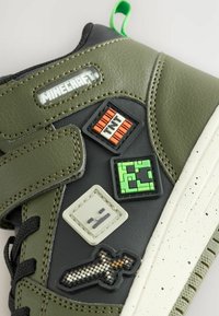 Baskets vertes et noires avec des patchs en caoutchouc sur le thème de Minecraft, incluant "MINECRAFT," un bloc TNT, un visage de Creeper, une épée pixelisée et des icônes de visage gris.
