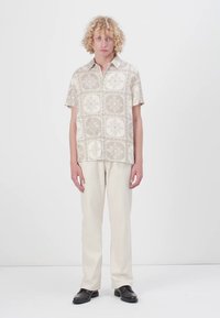 Kortermet mønstrete skjorte i beige med geometriske design, sammen med lysebeige bukser og svarte sko. Enkelt bakgrunn.