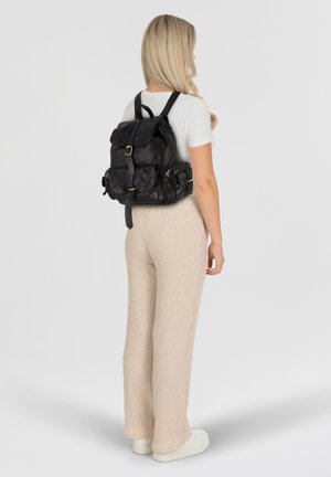 Mochila de cuero negro con solapa, herrajes dorados y bolsillos laterales. El modelo lleva pantalones de canalé crema y zapatos claros, de espaldas.