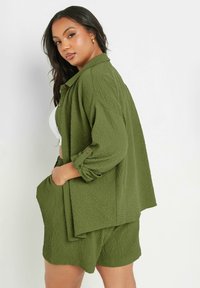 Completo in tessuto testurizzato verde oliva, composto da una camicia leggera con bottoni e pantaloni corti coordinati con tasche laterali. Taglio casual e rilassato.