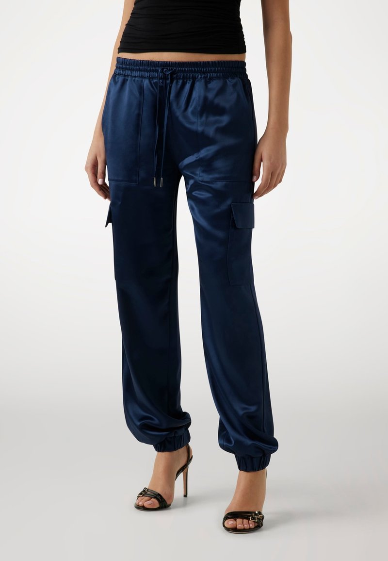 Marina satin cargo-byxor med elastisk midja, sidofickor och samlade anklar, tillsammans med svarta klackar med remmar.