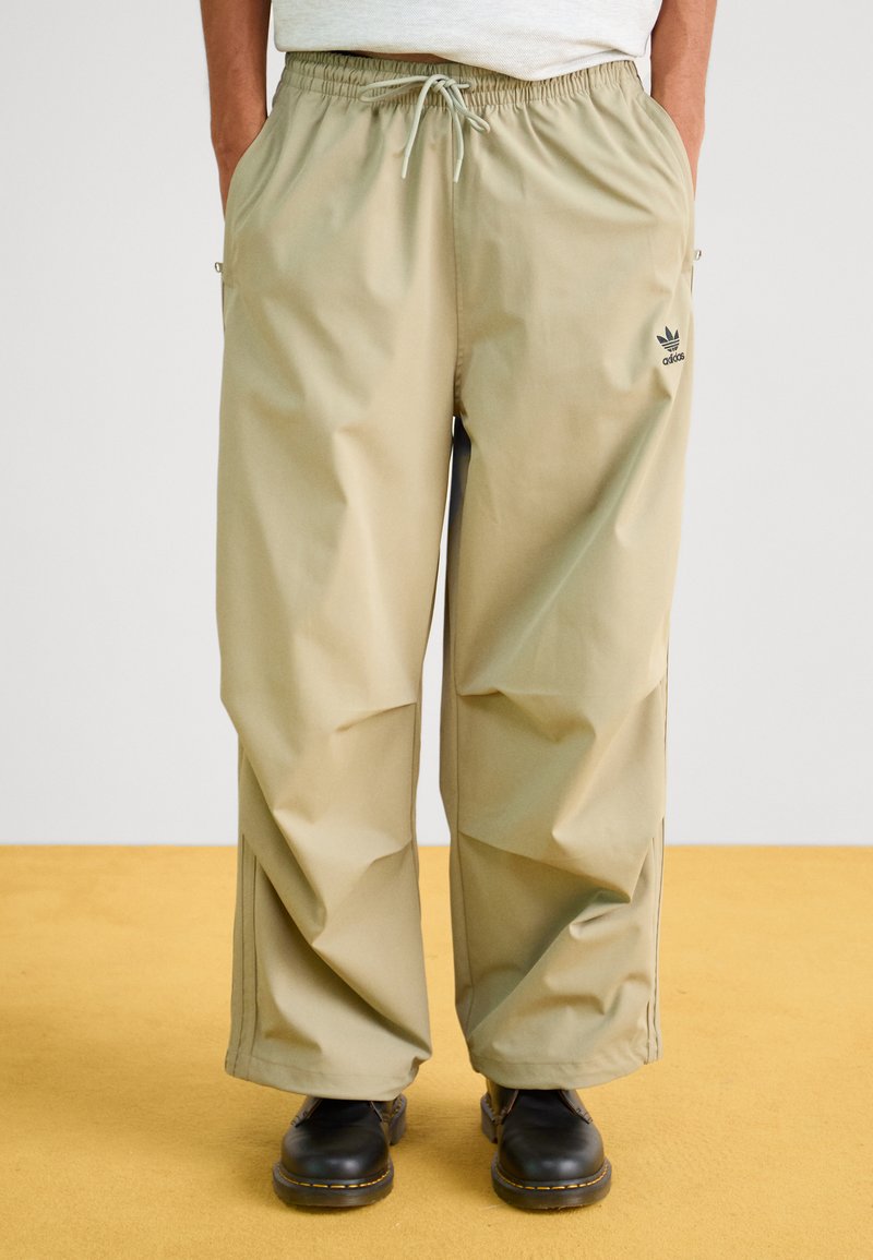 Μπεζ ελαφριά παντελόνια με ελαστική μέση, δύο πλαϊνές τσέπες, διακριτική λεπτομέρεια λογότυπου και άνετη γραμμή, συνδυασμένα με μαύρα παπούτσια.