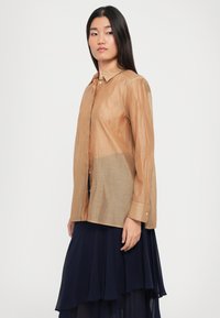 Femme avec de longs cheveux noirs, portant une chemise beige semi-transparente à boutons et une jupe bleu marine à couches, se tenant contre un fond clair.