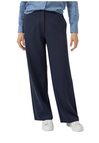 Marineblauwe wijde broek met plooien, gemaakt van een zachte stof, gecombineerd met witte sneakers voor een casual uitstraling. Details zijn minimaal en strak.