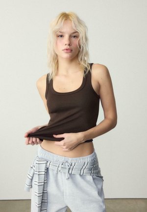 Junge Frau mit blondem Haar trägt ein braunes Tanktop und graue Jogginghosen, hält das Ende ihres Oberteils vor einem schlichten Hintergrund fest.