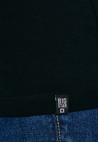 Zwart stof met een label "BIG STAR" genaaid vlak bij de zoom boven blauwe spijkerbroek met zichtbare stiknaden.