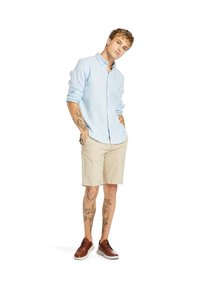 Camicia azzurro chiaro con bottoni, pantaloni corti beige, scarpe marroni. Tattoo prominenti sulle braccia. Modello in piedi con le mani nelle tasche, leggermente inclinato.