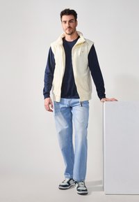 Lys cremefarvet ærmeløs zip-up vest over en mørkeblå langærmet skjorte, parret med lyseblå jeans og sort-hvide sneakers.