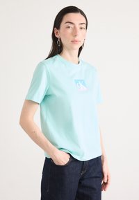 Bledomodrý tričko z bavlny s námorníckym štvorcovým logom v bielych a modrých odtieňoch. Krátke rukávy a voľný strih, skombinované s tmavými džínsami.