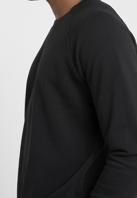Sudadera negra de manga larga con cuello redondo, puños de canalé, textura suave y diseño ajustado, con costura visible en el hombro.