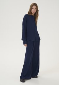 Camicia button-up blu navy con maniche lunghe, abbinata a pantaloni blu larghi. Tessuto morbido, design minimale e silhouette rilassata.