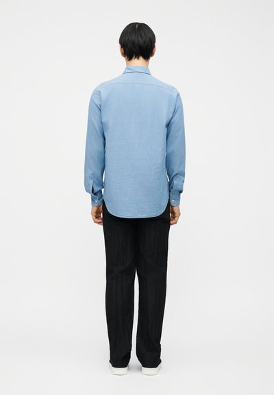 Άτομο που στέκεται με την πλάτη προς τα εμάς, φορώντας γαλάζιο μακρυμάνικο πουκάμισο, μαύρο παντελόνι και λευκά παπούτσια, μπροστά σε απλό λευκό φόντο.