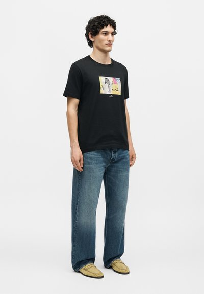 Άνδρας που φοράει μαύρο κοντομάνικο T-shirt με πολύχρωμο σχέδιο ζέβρας, μπλε τζιν παντελόνι με φαρδιά πόδια και μπεζ μοκασίνια, στέκεται σε λευκό φόντο.