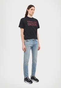 Svart t-skjorte med en rød "Versace Jeans Couture" grafikk, kombinert med lyseblå slim-fit jeans og svarte sneakers med hvite detaljer.