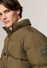 Doudoune vert olive avec col montant, fermeture éclair et patch logo sur l'épaule gauche. Texture lisse et design matelassé pour l'isolation.