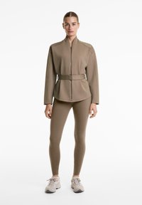 Egy nő taupe színű, zipzáros kabátot visel övvel és hozzáillő leggingset, amelyet világos bézs sportcipővel párosít, miközben egy fehér háttér előtt áll.