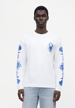 Homme à la peau foncée portant une chemise blanche à manches longues avec des motifs bleus de pieuvre et de coquillages sur les manches et la poitrine, associée à un jean bleu clair.