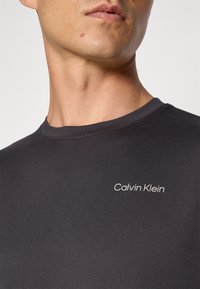 Juodas trumpomis rankovėmis marškinėliai, pagaminti iš lygios medžiagos, su apvalia iškirpte ir mažu baltu "Calvin Klein" logotipu ant krūtinės.