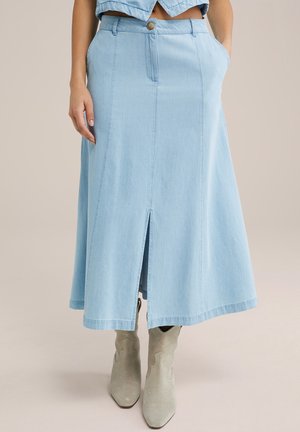 Femme portant une jupe midi en denim bleu clair avec une fente à l'avant et des bottes de cowboy beiges, debout avec une main dans la poche.