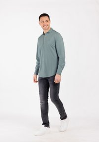 Uomo che cammina, indossando una camicia verde chiaro con bottoni, jeans grigio scuro e scarpe da ginnastica bianche, sorridendo e guardando di lato.