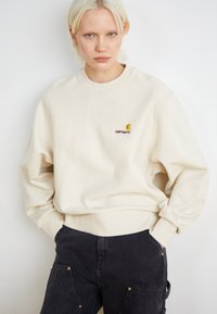 Beige sweatshirt med ribbet halsudskæring og manchetter, prydet med et lille logo i gul og bordeaux. Parret med mørke denim cargo bukser.