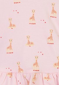 Tessuto rosa chiaro con un motivo ripetuto di giraffe con macchie rosse, insieme a piccole stelle rosse e un orlo arricciato nella parte inferiore.