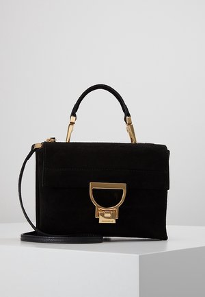 Sac à main en daim noir avec des accessoires dorés, une poignée supérieure et une bandoulière amovible, présenté sur une surface blanche.