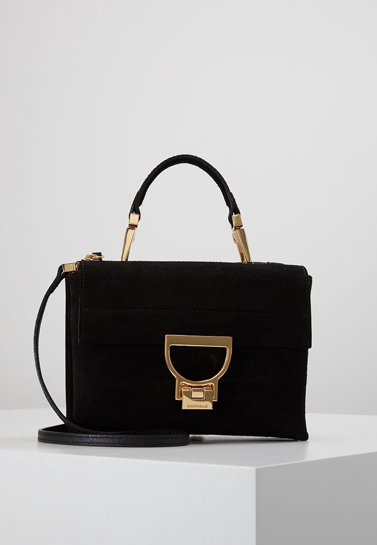 Sac à main en daim noir avec des accessoires dorés, une poignée supérieure et une bandoulière amovible, présenté sur une surface blanche.