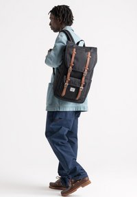 Schwarzer Rucksack mit braunen Lederriemen und Metallschnallen, ausgestattet mit einem großen Klappdeckel und einem Logo-Patch auf der Vorderseite. Getragen von einer Person in legerer Kleidung.